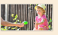 Ein kleines Kind in rosa Kleidung und gelbem Sonnenhut sitzt draußen auf einem Stein oder einer Mauer und erhält von einer erwachsenen Hand ein grünes Spielzeug.