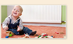 Ein Kleinkind sitzt auf einem Teppich vor einem Heizkörper und spielt fröhlich mit bunten Holzklötzen. Das Kind trägt eine Latzhose und ein kariertes Hemd.